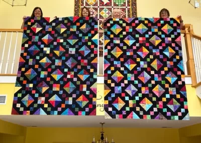 Two colorful patchwork quilts held up against a yellow wall with a staircase.