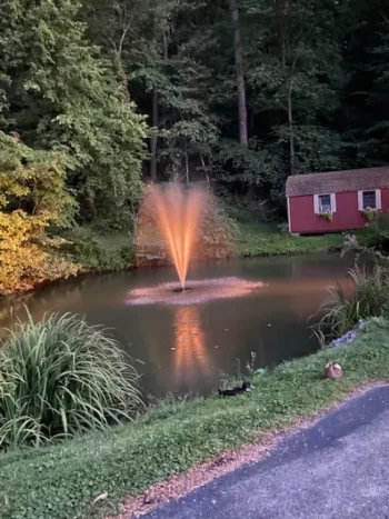 A serene pond with a fountain at dusk, surrounded by trees and a red shed in the background.