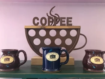 A decorative coffee cup-shaped sign with the text "COFFEE" above three ceramic mugs on a shelf.